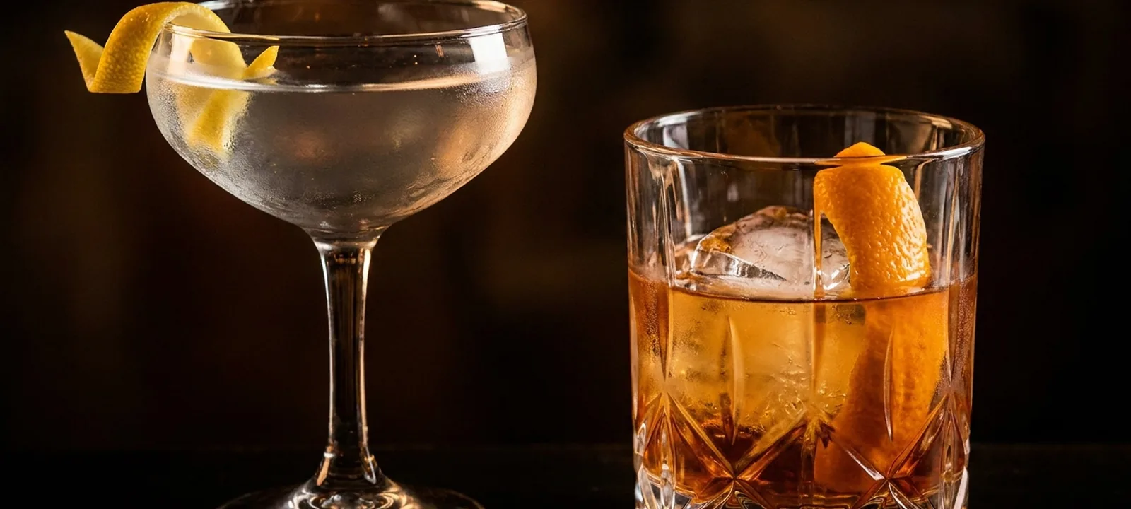 Zwei elegante Cocktails an einer dunklen Bar, einer mit Zitronenzeste im Coupé-Glas, der andere mit Eis und Orangenscheibe